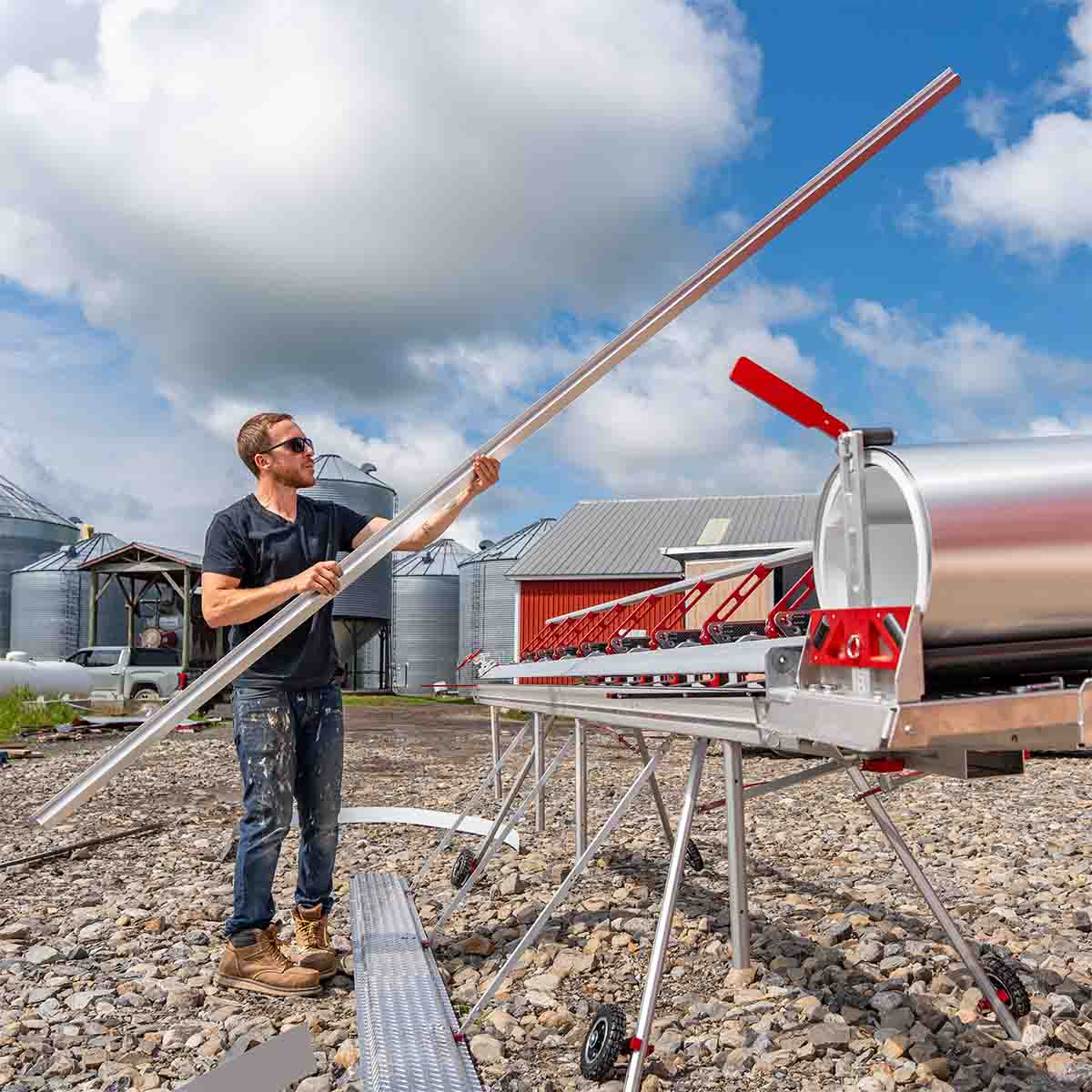 Man holding and looking at a 14 foot siding sheet metal piece he just bent on an Innovatools bending machine, with a red barn and farm silos in the background.