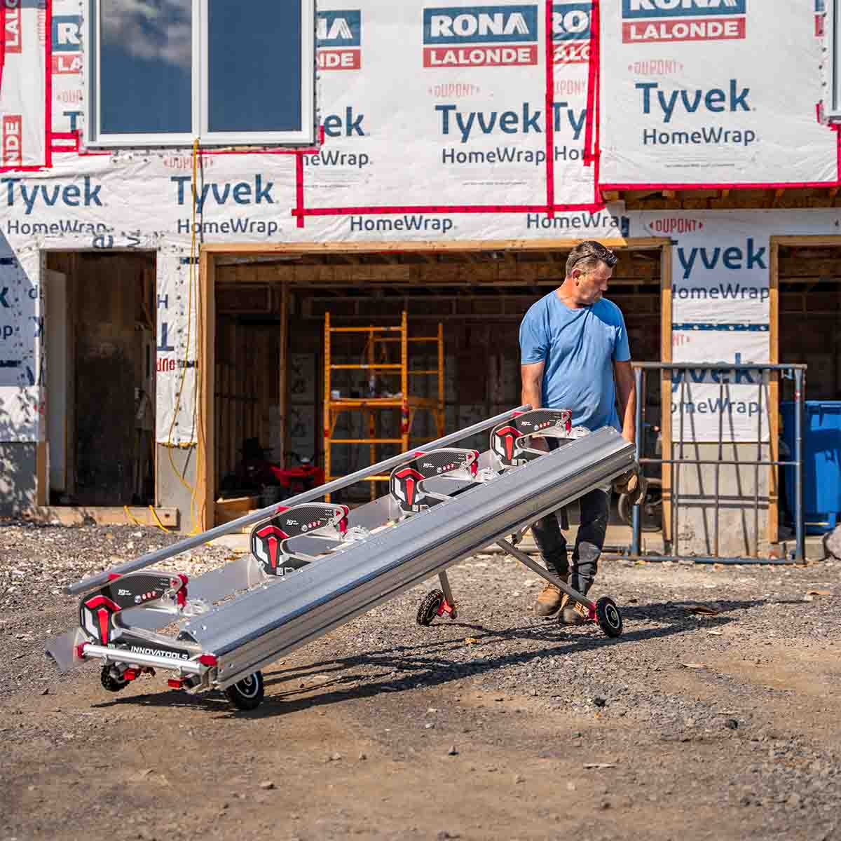 A man carries an Innovatools bending machine on an Innovatools collapsible stand with four wheels, in front of a house under construction. The two far wheels of the stand are retracted, while the other two wheels are extended.
